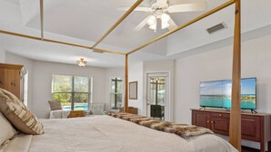 Une chambre spacieuse avec un grand lit, un ventilateur de plafond, une télévision et une vue sur la piscine par les fenêtres.
