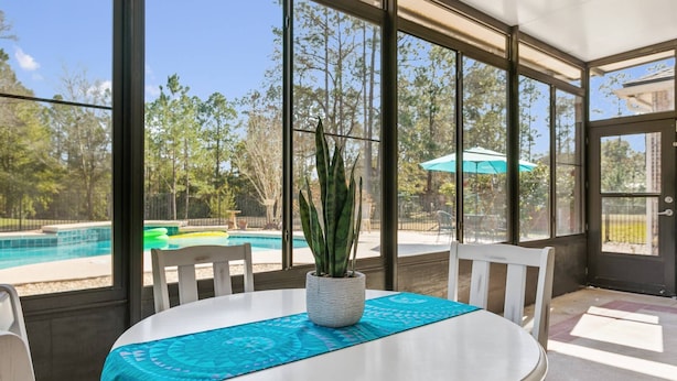 Une table ronde avec un plateau blanc et une nappe bleue à motifs, une plante en pot et des chaises blanches dans une pièce dotée de grandes fenêtres donnant sur une piscine et un espace de détente extérieur.