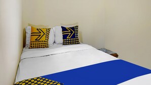 Een bed met een wit-blauwe sprei, twee kussens met gele en blauwe patronen, en een houten nachtkastje met een lamp.
