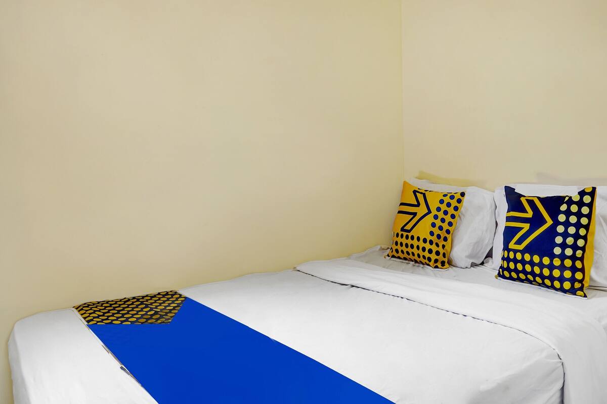 Una cama con ropa de cama blanca y un detalle en azul, dos almohadas con diseños azules y amarillos, y una pared lisa.