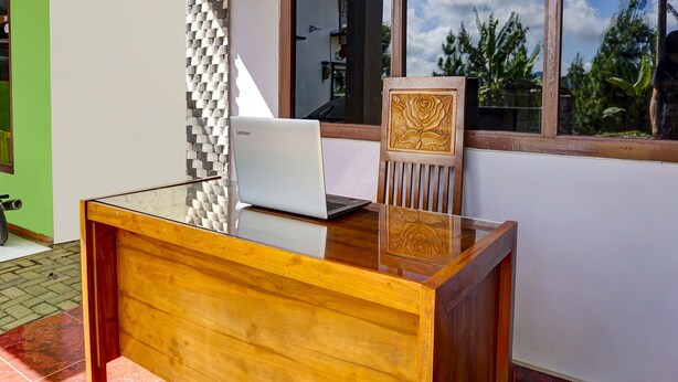Een houten bureau met een laptop, een stoel met een houtsnijmotief en een raam met uitzicht op groen.