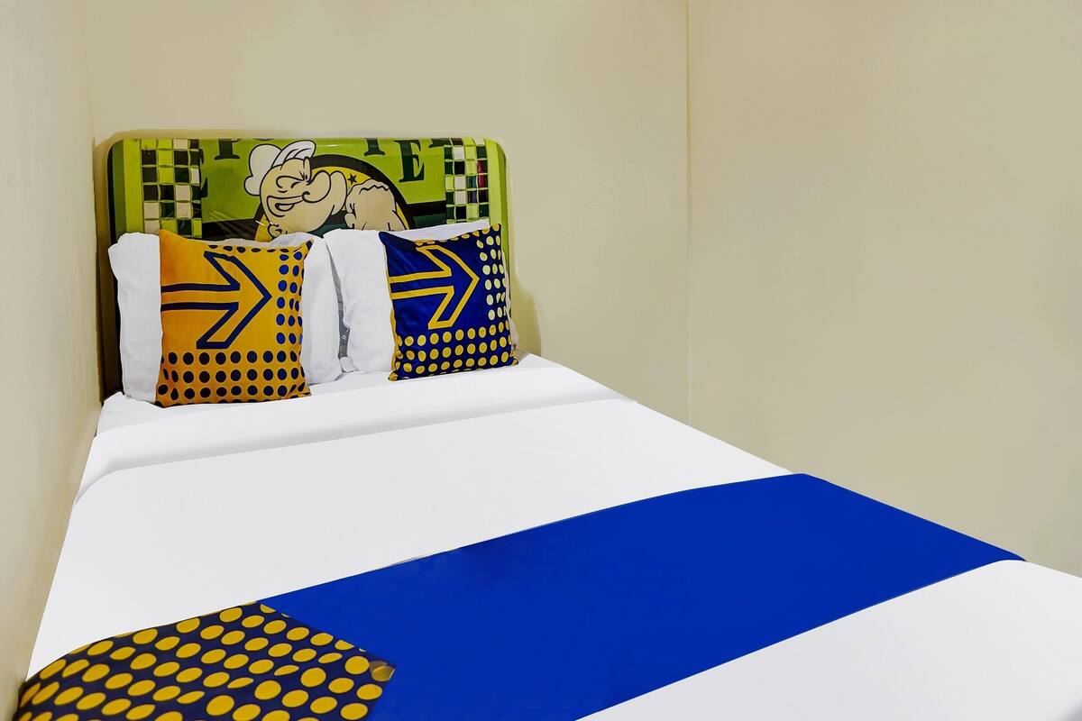 Una cama con una colcha blanca y azul, dos almohadas con diseños amarillos y azules, y un cabecero verde con un personaje de dibujos animados.