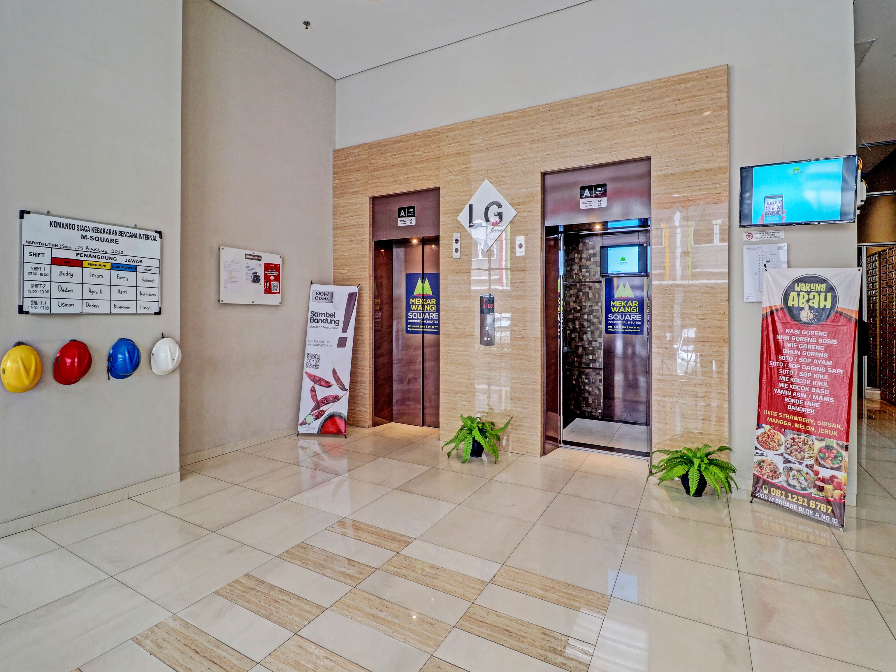 A modern lobby with a tiled floor, a reception desk, and an elevator. There are safety helmets on the wall, a signboard, and a digital screen displaying a menu.