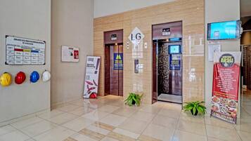 A modern lobby with a tiled floor, a reception desk, and an elevator. There are safety helmets on the wall, a signboard, and a digital screen displaying a menu.