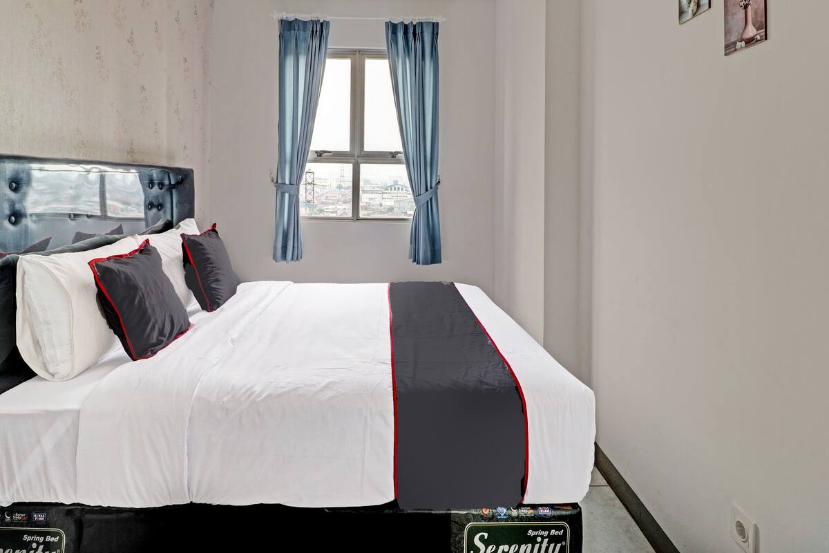 Una cama bien hecha con cabecera, ropa de cama blanca con detalles en rojo y negro, y una ventana con cortinas azules.