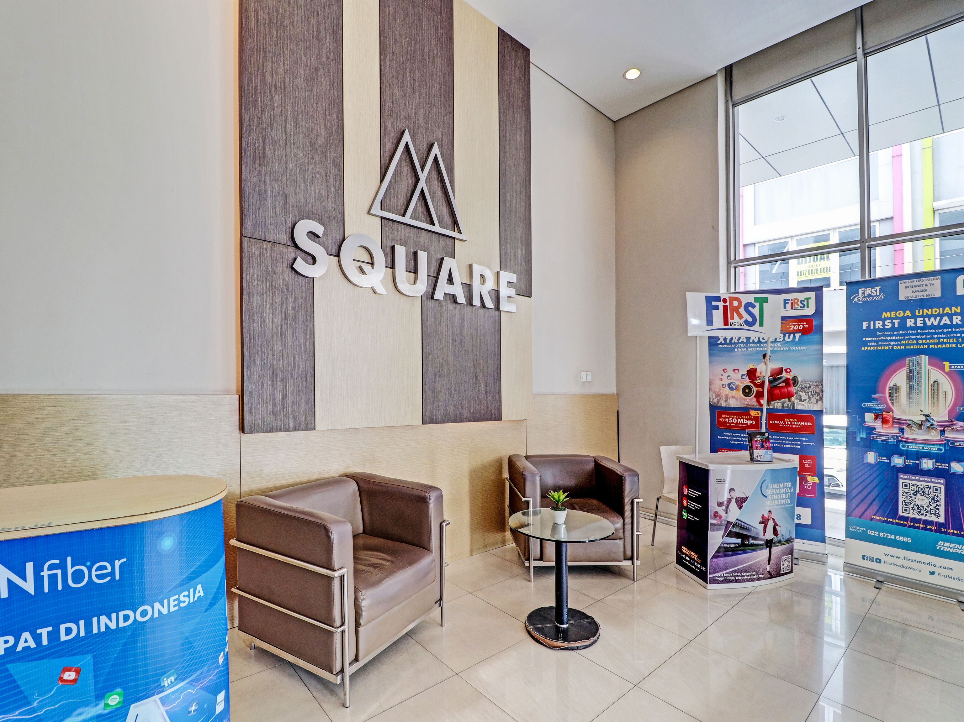 A modern lobby with a 'SQUARE' sign, leather seating, a glass table, and a 'First' advertisement.