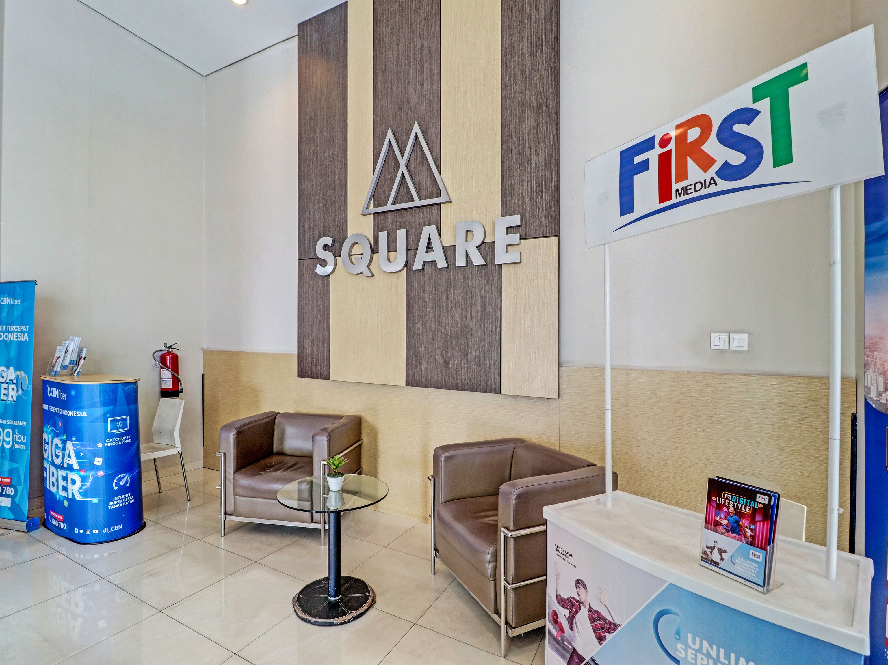 A lobby with a reception desk, two leather sofas, a glass table with a plant, and a sign for 'First Media'.
