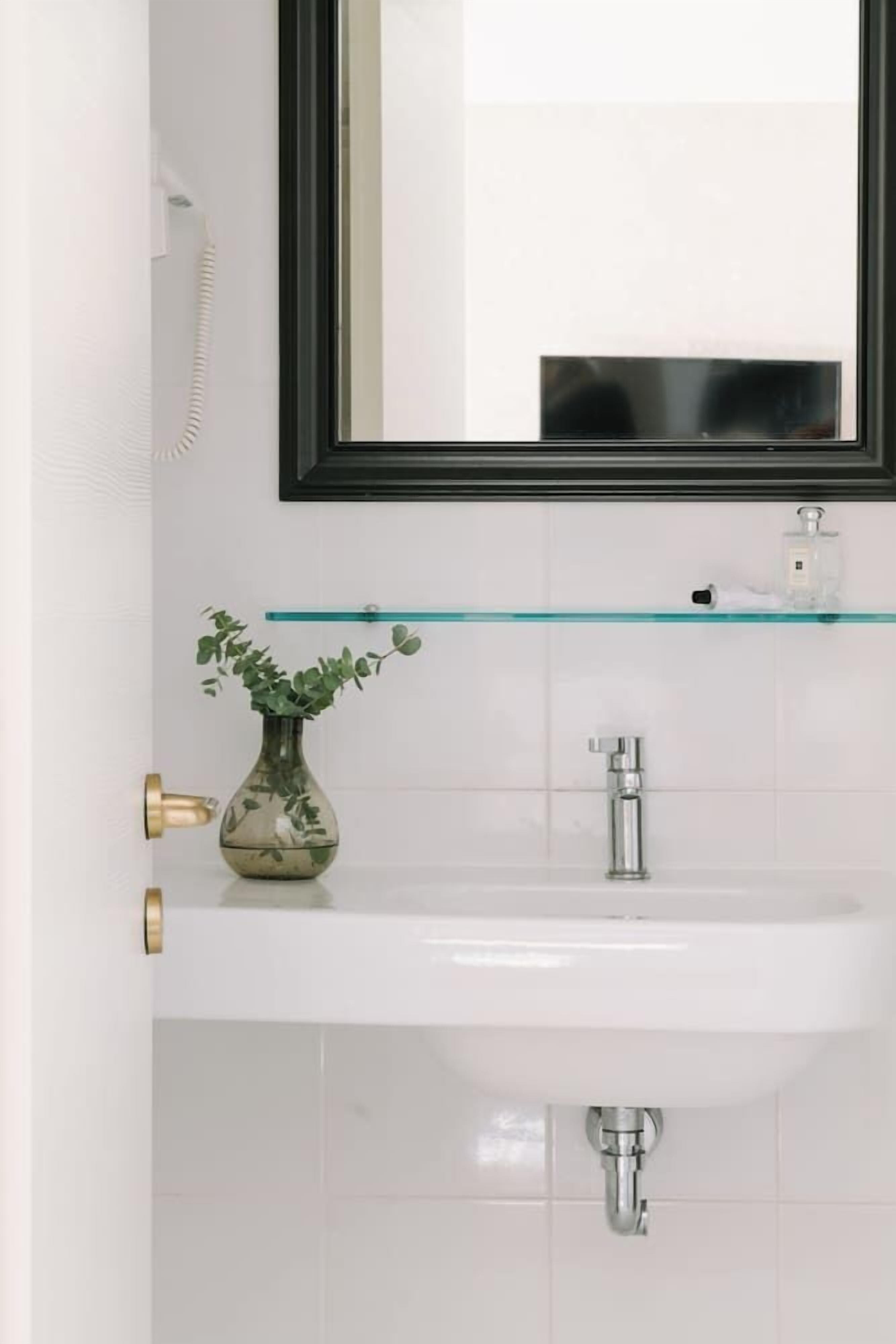 Un baño con un lavamanos blanco, un estante de vidrio arriba, un florero con una planta y un espejo con marco negro.