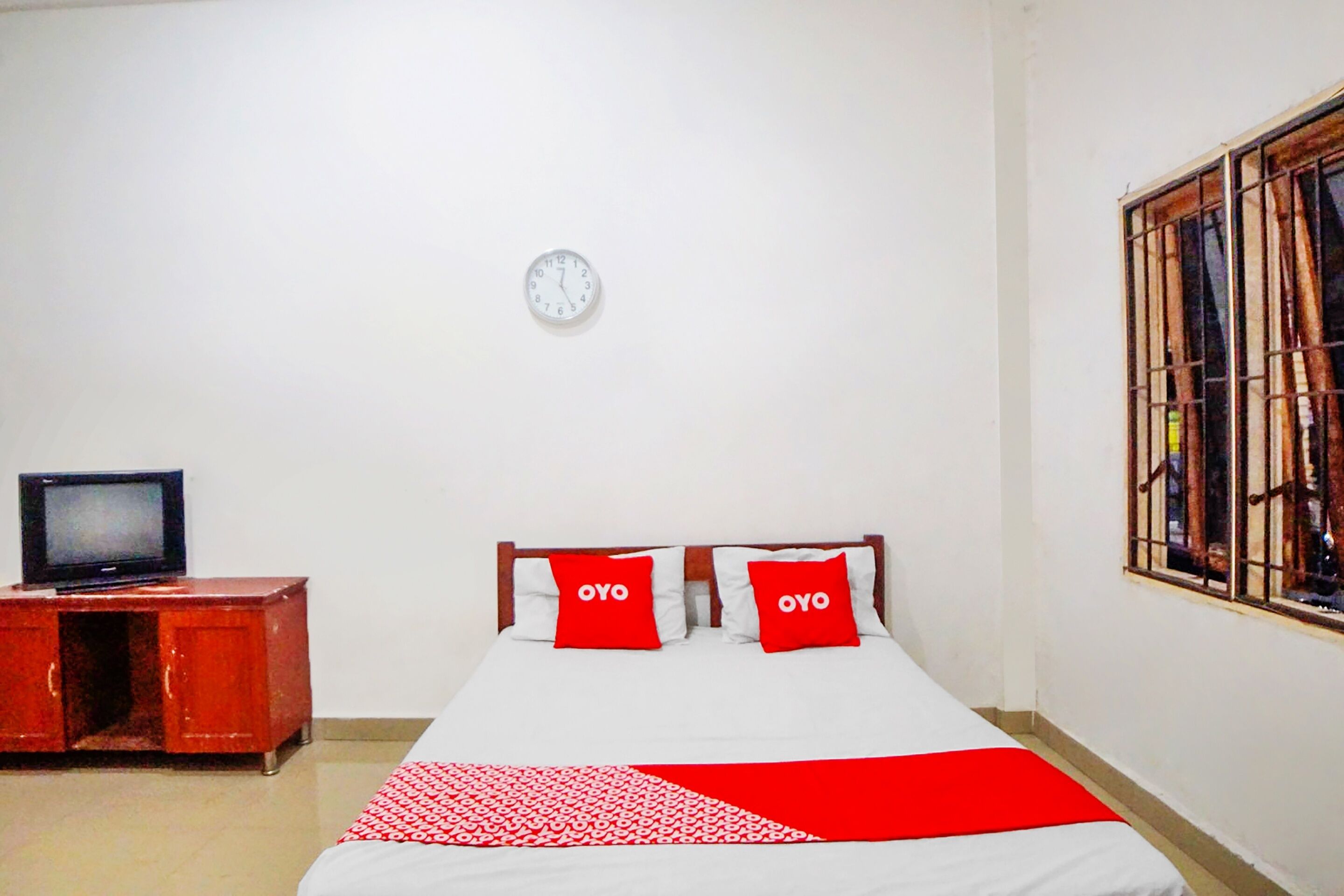 A room with a bed, two red pillows, a clock on the wall, a television on a wooden cabinet, and a window with bars.