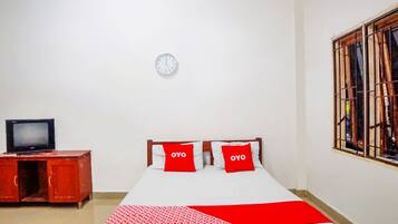 A room with a bed, two red pillows, a clock on the wall, a television on a wooden cabinet, and a window with bars.
