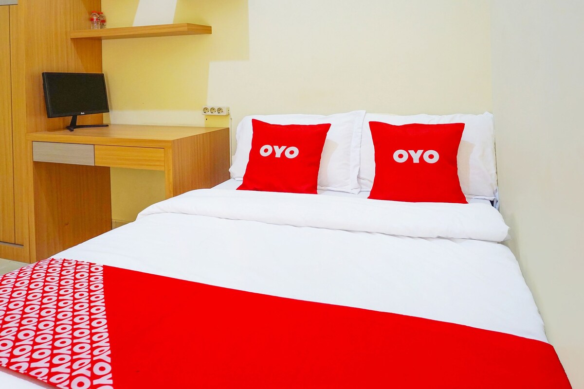 Et hotelværelse med en seng, to røde puder med teksten "OYO", et hvidt sengetæppe, et rødt og hvidt mønstret tæppe, et træskrivebord med en computerskærm og en hylde med småting.