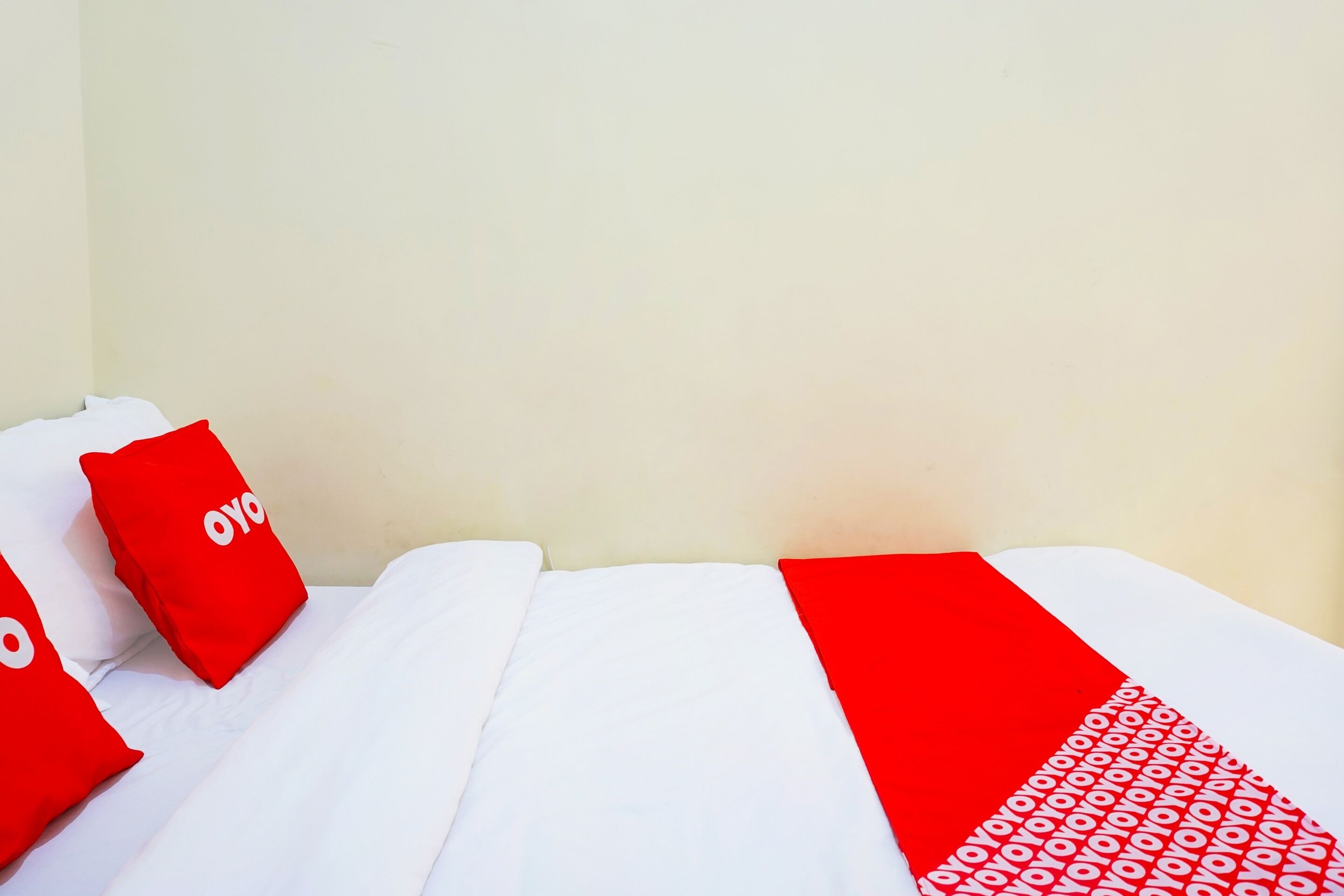 A bed with white bedding and red pillows, a red blanket with a pattern, and a plain wall in the background.