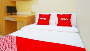 A hotel room with a bed, two red pillows with "OYO" text, a white bedspread, a red and white patterned blanket, a wooden desk with a computer monitor, and a shelf with small items. - Flagship 90723 Berlima Guest House Syariah (Malang)