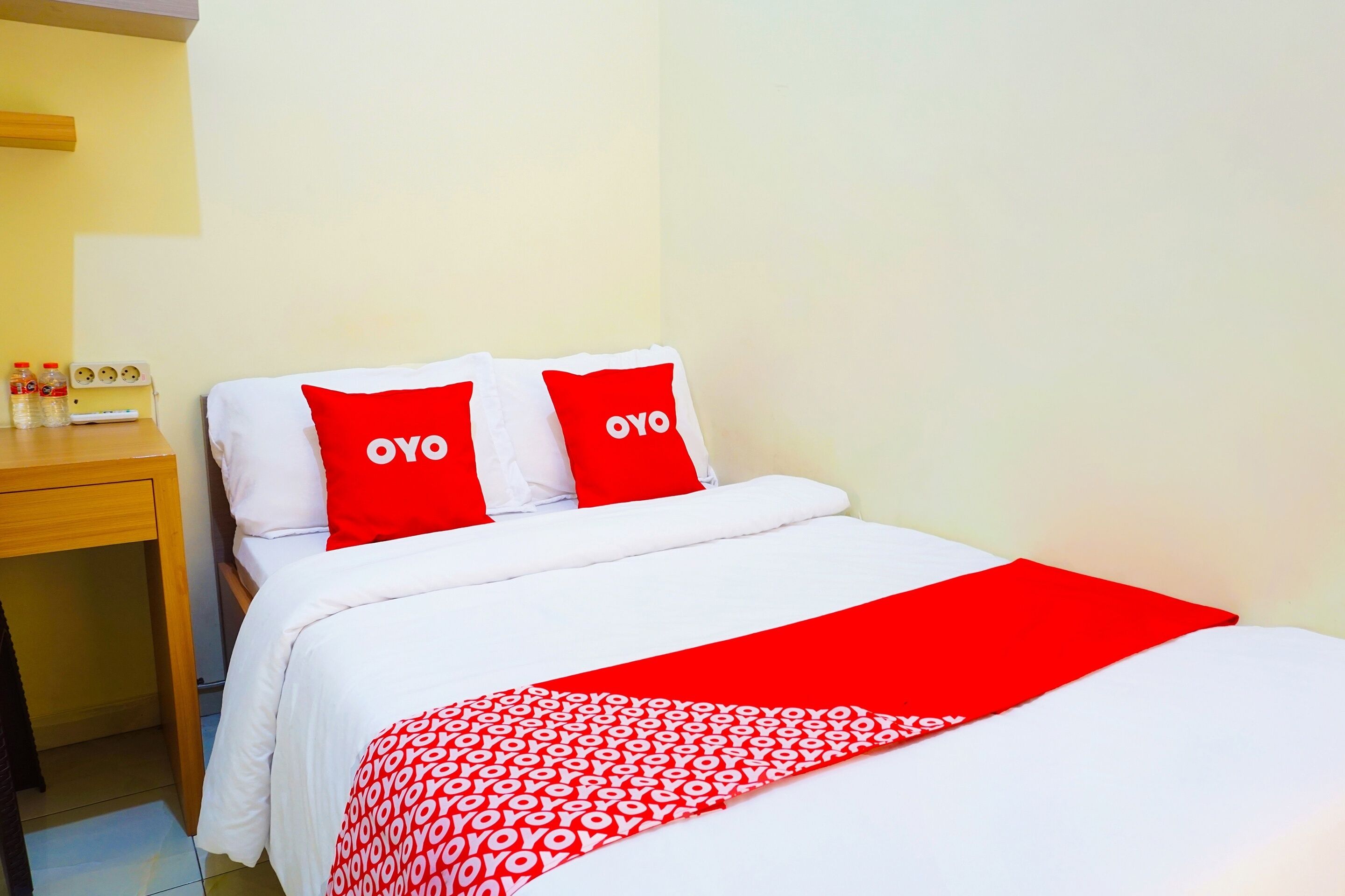 A hotel room with a bed, two red pillows with "OYO" branding, a wooden nightstand, and a red and white patterned throw blanket.