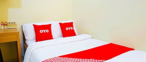 A hotel room with a bed, two red pillows with "OYO" branding, a wooden nightstand, and a red and white patterned throw blanket.