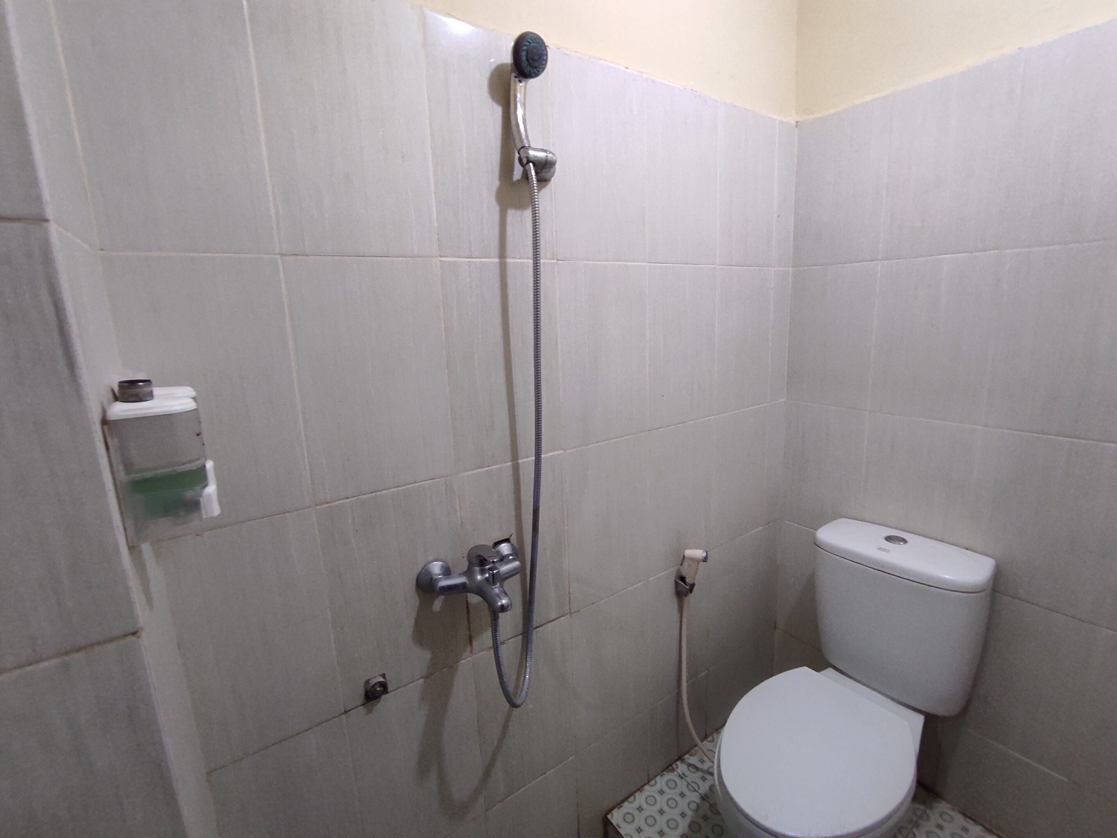 A small, tiled bathroom with a toilet, a shower, and a wall-mounted soap dispenser.