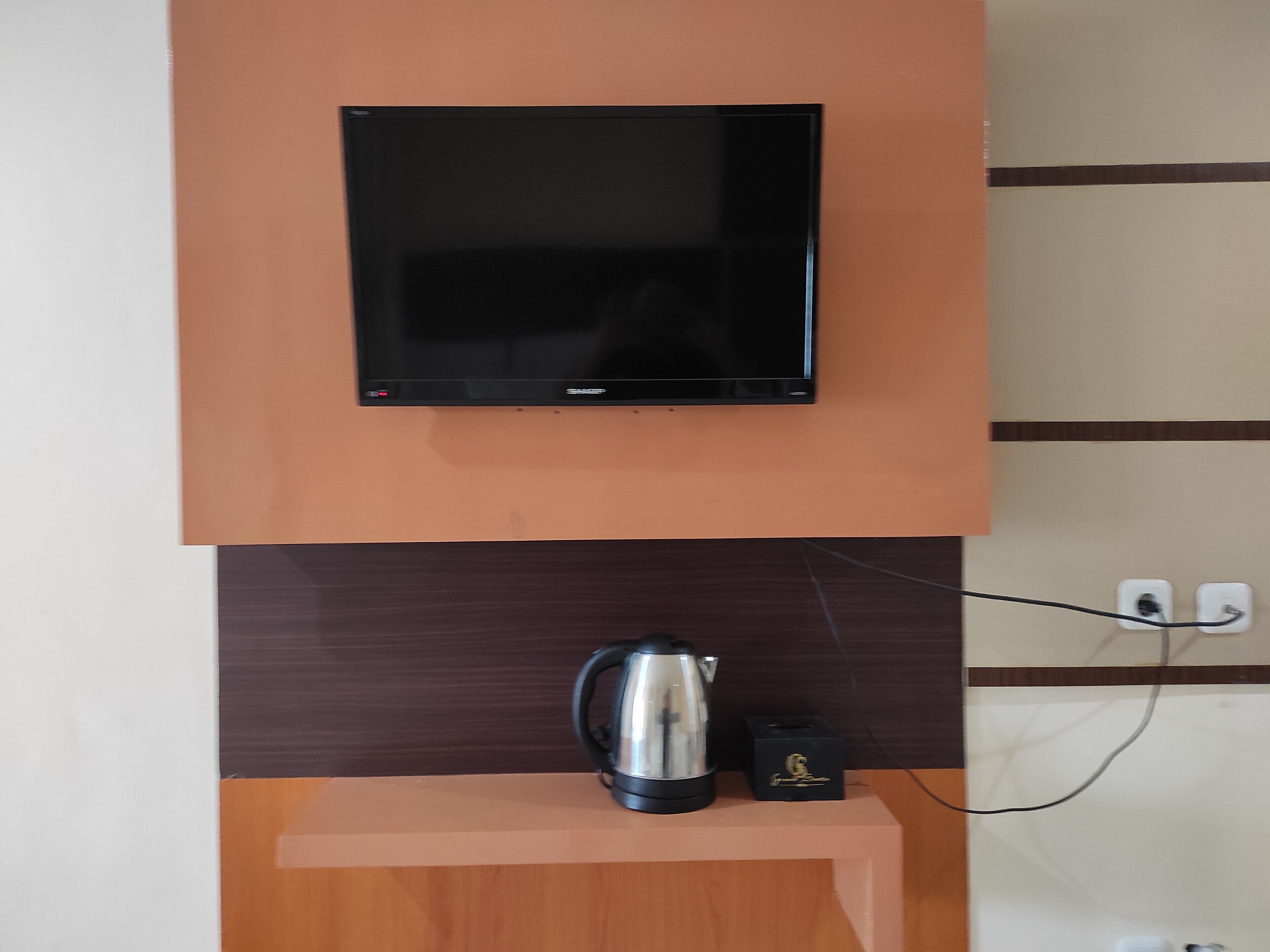 A flat-screen TV mounted on a wall, a kettle on a wooden shelf below, and a power outlet with a cable.