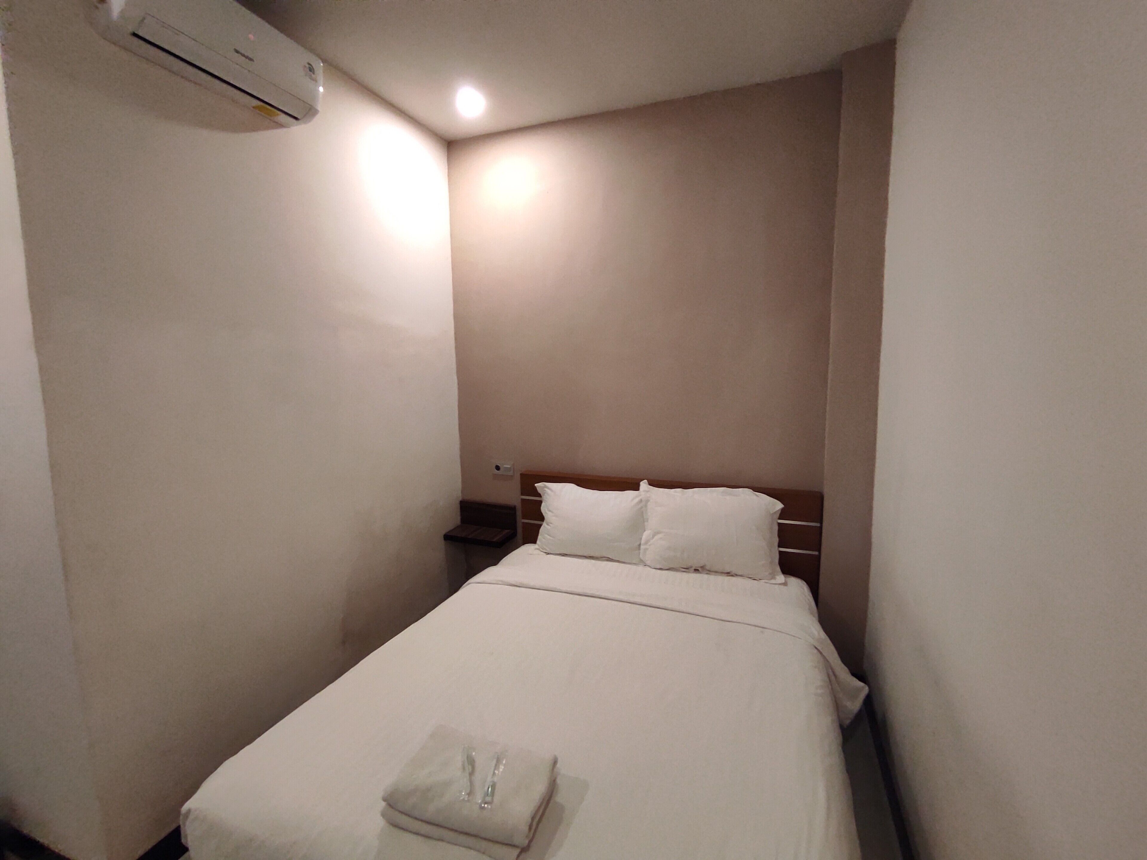 A small, neatly made bed with white linens and pillows in a compact room with plain walls and a single bedside table.