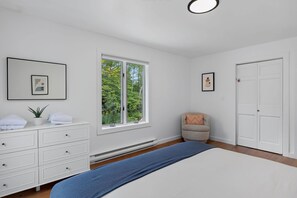 A bedroom with a bed, a dresser, a chair, a window with a view of trees, and a door.