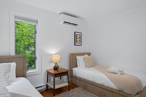 A bedroom with a bed, bedside table, lamp, air conditioning unit, and a window with a view of greenery.
