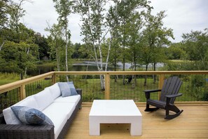 A deck with a sofa, a table, and a rocking chair, overlooking a lake with trees.