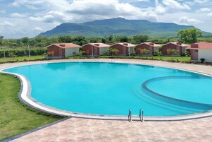 Piscina com borda curva, rodeada por um deck pavimentado. Ao fundo, vários edifícios pequenos com telhados vermelhos, provavelmente parte de um resort ou hotel.