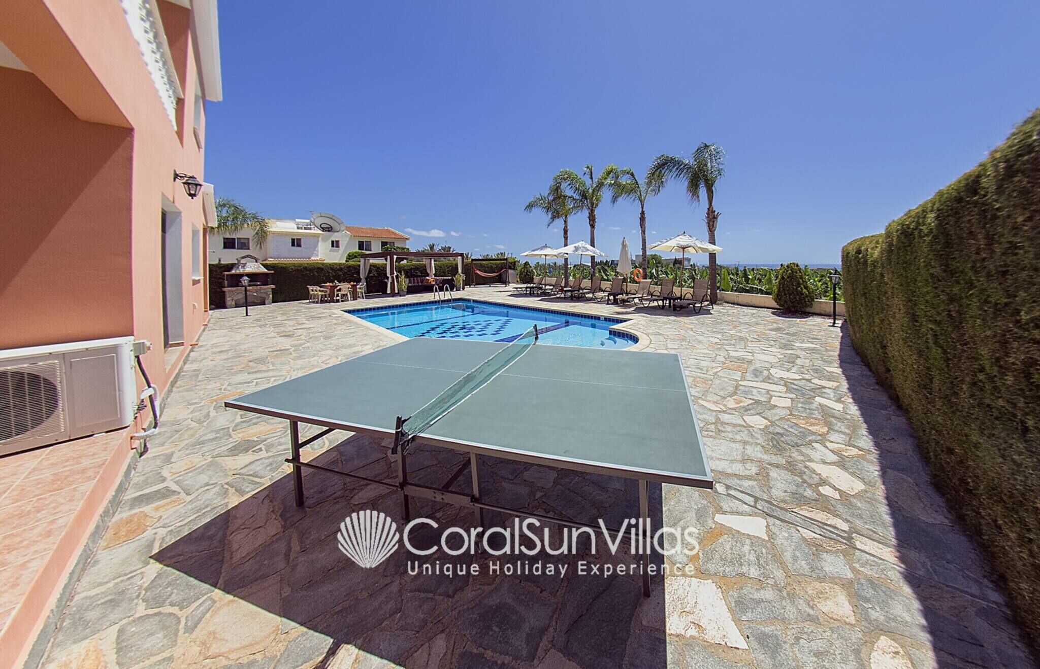 A swimming pool with lounge chairs and umbrellas, a ping pong table, and a well-maintained hedge.
