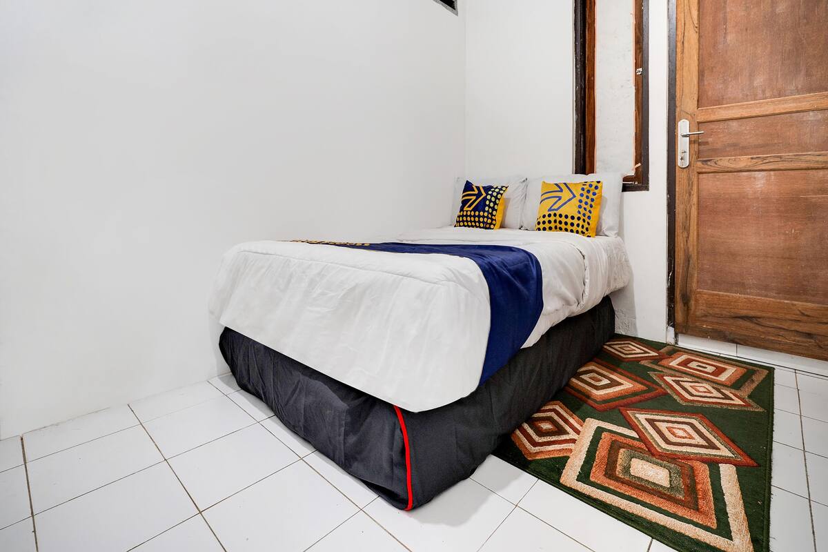 A neatly made bed with a white and blue bedspread, two patterned pillows, and a dark skirted bed base. A wooden door is visible to the right, and a patterned rug is on the floor.