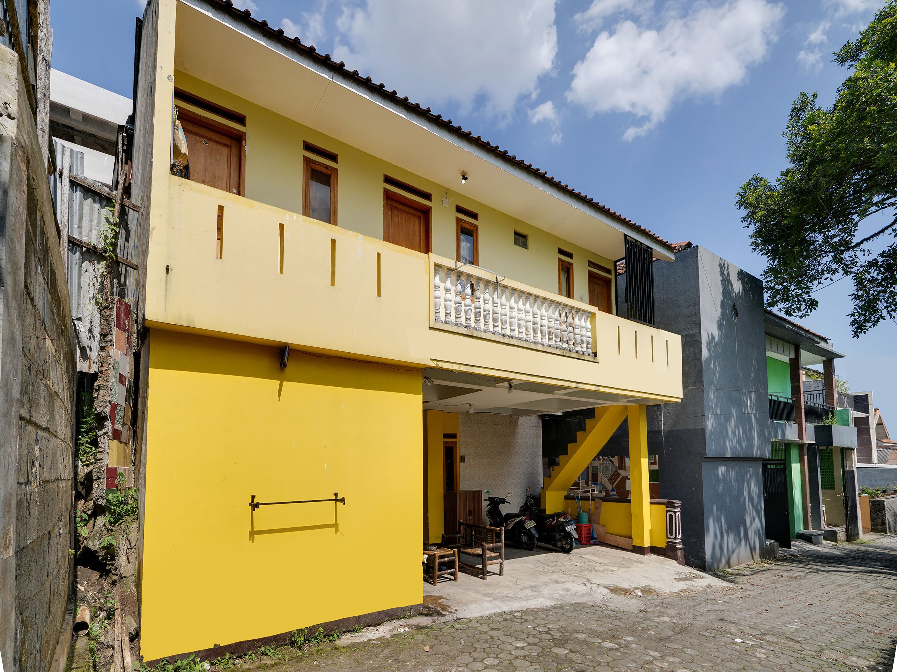 A yellow building with a balcony, a scooter parked in front, and a narrow alleyway.