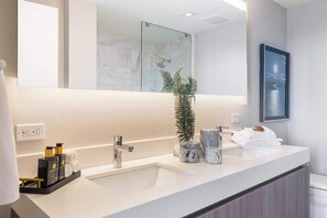 A modern bathroom with a large mirror, a sink, and a shower. There is a plant on the counter and towels stacked on the side. - Luxury Beach Condo | Pool, Gym & Balcony (Fort Lauderdale)