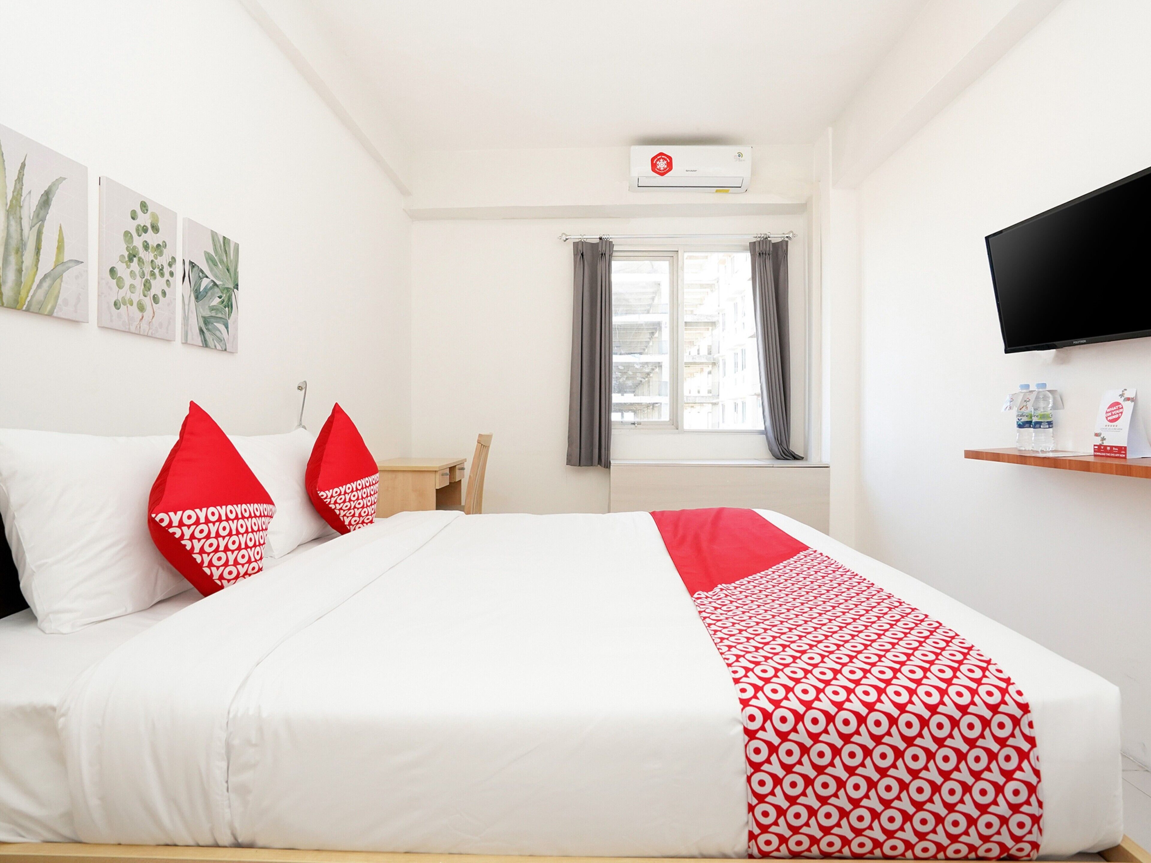 A hotel room with a bed, a desk, a chair, a window with curtains, a mounted TV, and artwork on the wall.