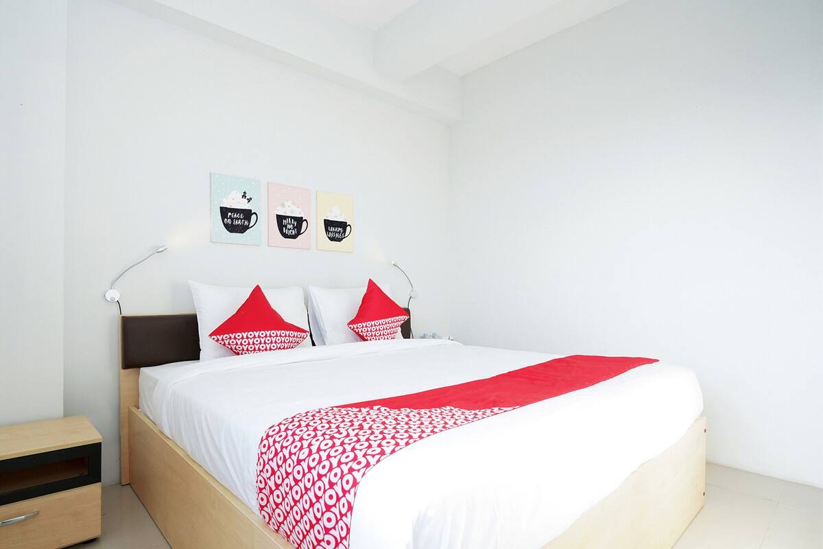 A neatly made bed with a white and red bedspread, two red pillows, and a wooden nightstand with a drawer.
