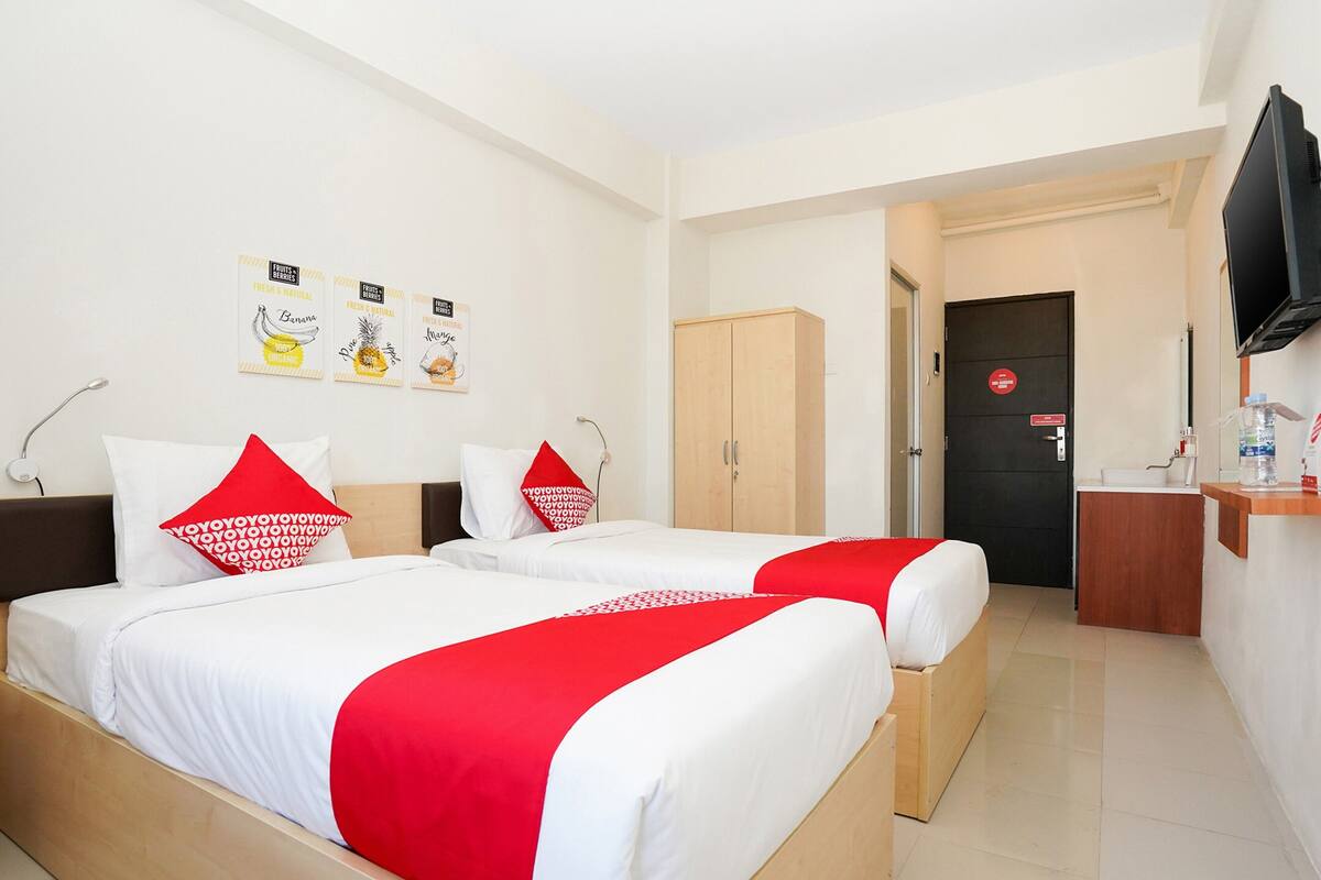 A hotel room with two beds, a wooden wardrobe, a desk with a TV, and a bathroom door.