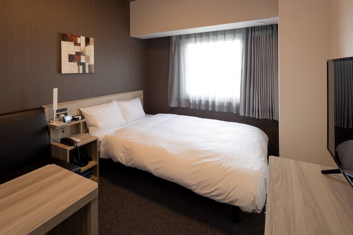 A hotel room with a large bed, a desk, a chair, a bedside table, a lamp, and a TV.