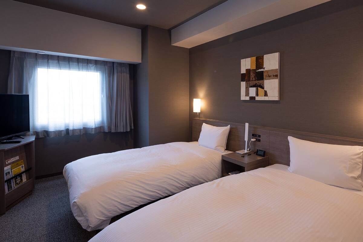 A hotel room with two beds, a TV, a desk, and a window with curtains.