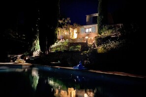 A nighttime view of a pool area with a stone wall and a building in the background. - Beautiful villa near Village des Vans (SAINT GENEST DE BEAUZON)