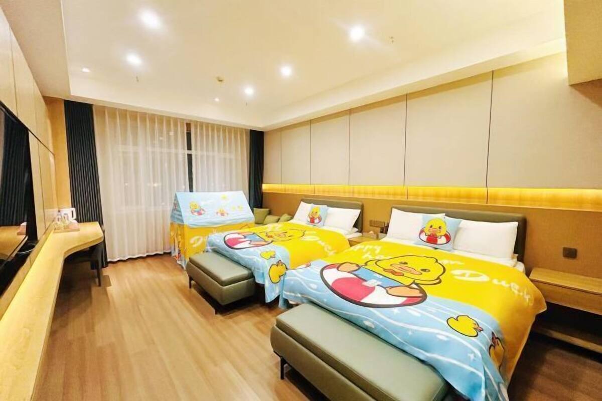 A hotel room with two beds, each with a cartoon-themed duvet cover, a wooden bench, a desk, and a large window with curtains.
