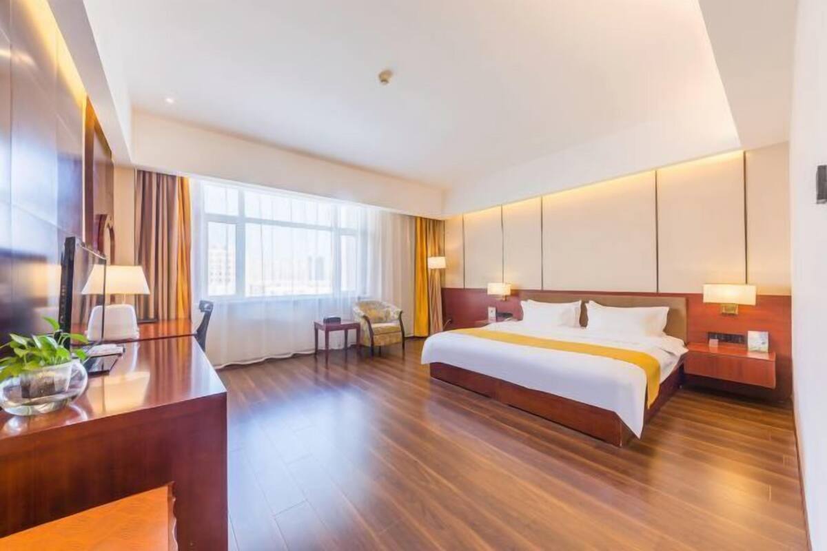 A hotel room with a large bed, a desk with a lamp, a chair, and a window with curtains.