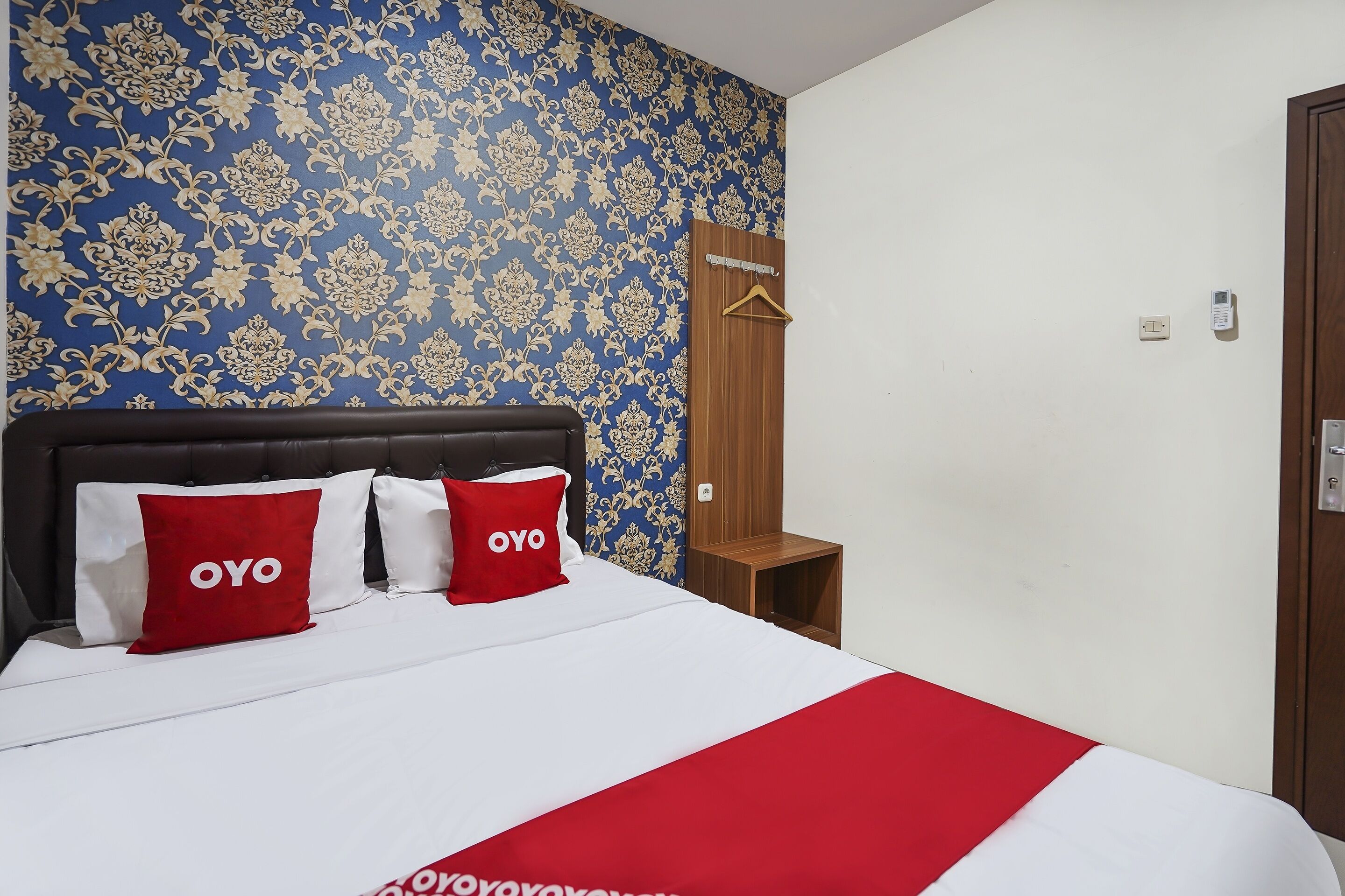 A hotel room with a bed, two red pillows with "OYO" branding, a wooden nightstand, and a wardrobe with a hanger.