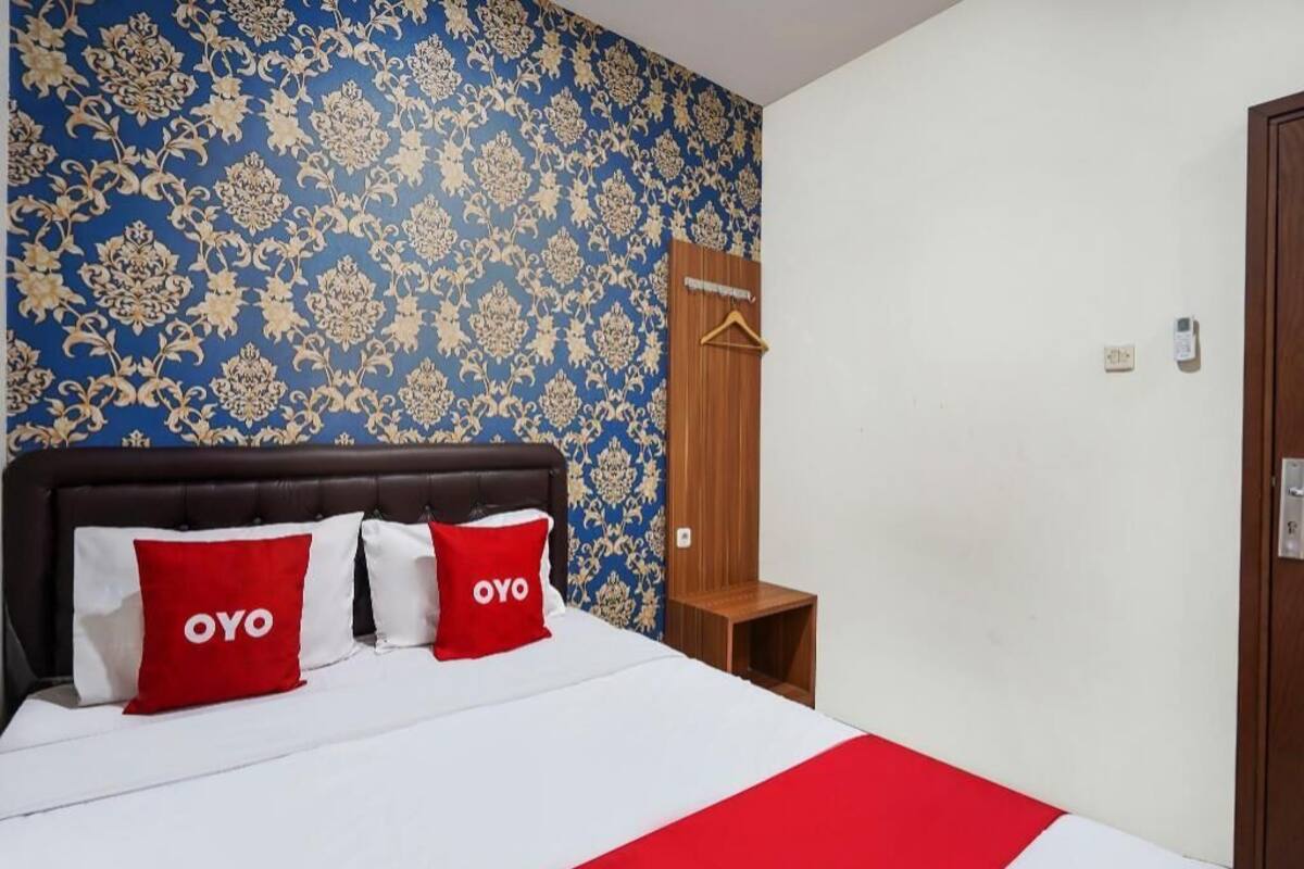 A hotel room with a bed, a wooden headboard, a blue patterned wall, and a red and white bedspread.