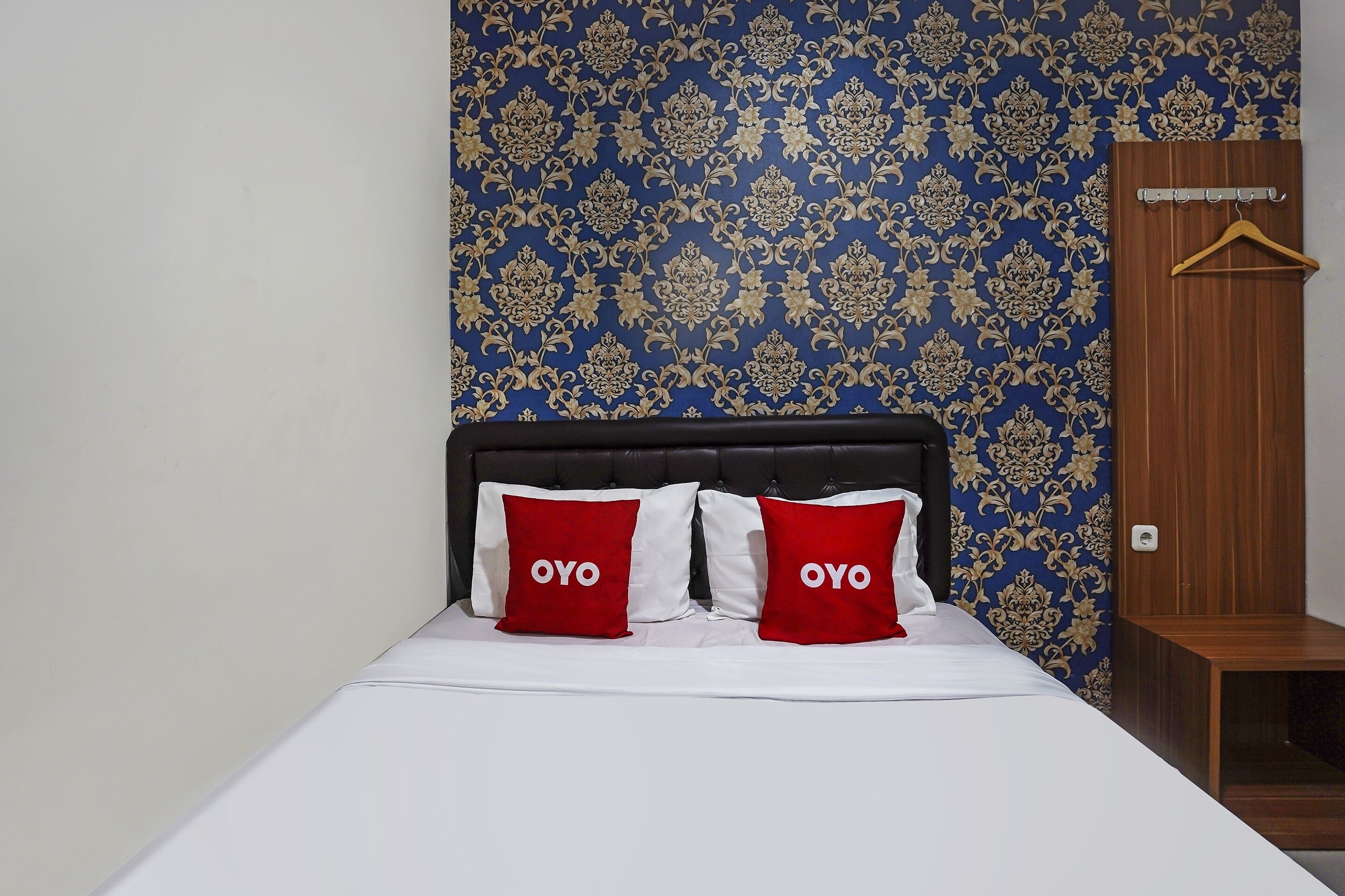 A bed with two red pillows labeled "OYO" and a wooden wardrobe with a hanging hook.
