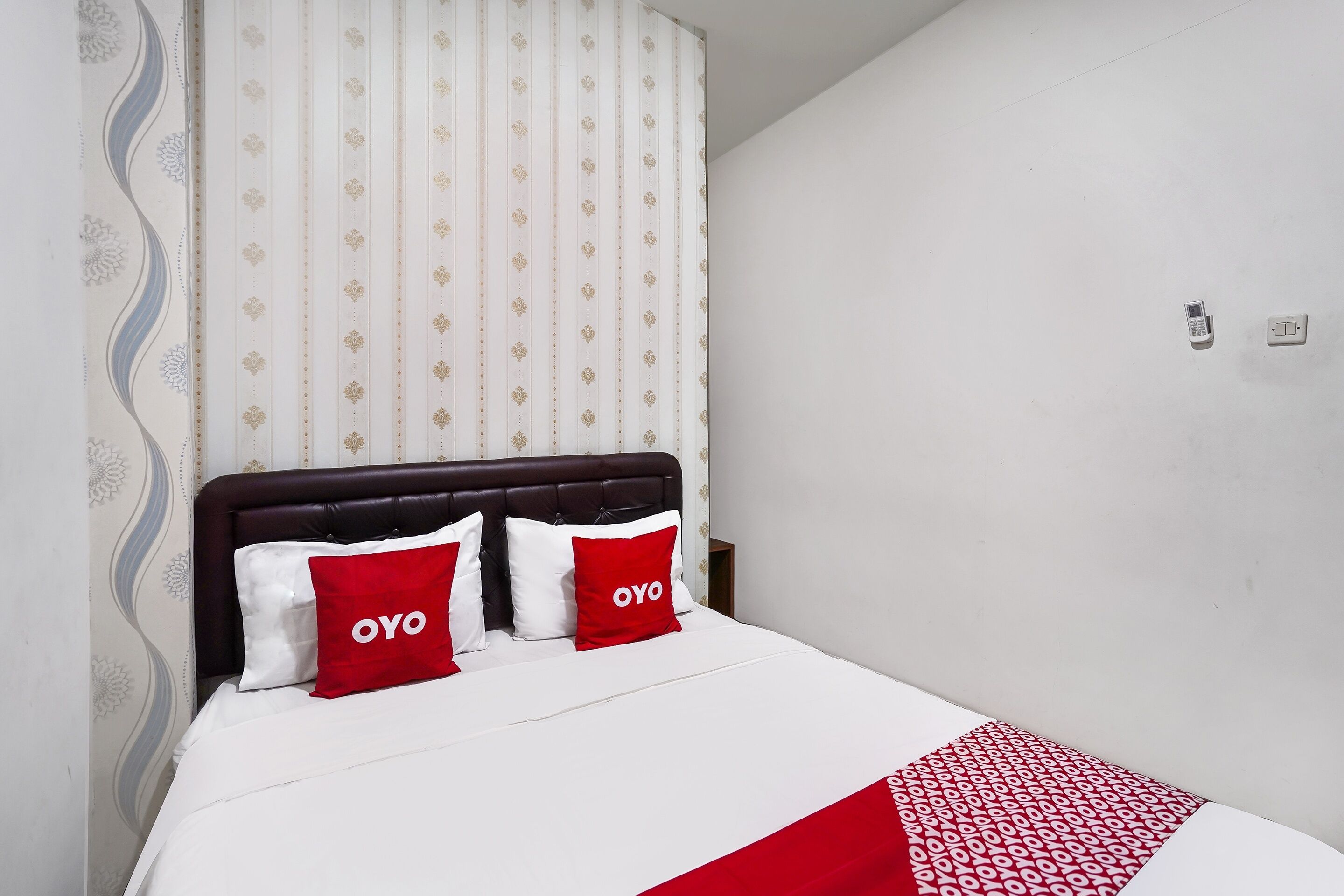 Una cama con colcha blanca y roja, dos almohadas rojas con la palabra "OYO" y un cabecero de madera oscura, además de una cortina blanca con estampado floral.