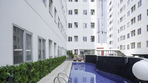 A modern indoor swimming pool with a tiled floor, surrounded by a low hedge and a building with multiple windows and balconies.