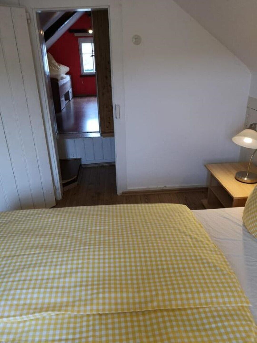 A bedroom with a checkered bedspread, a bedside table with a lamp, and a view of a red room through an open door.