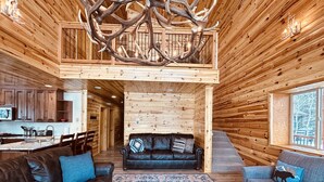 A log cabin interior with a central seating area, a kitchen, and a dining space.