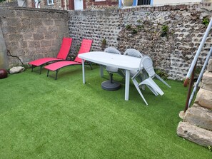 Un patio con una mesa blanca y sillas rojas, rodeado por un muro de piedra y césped artificial.