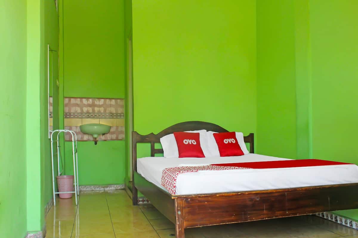 Un lit simple avec un cadre en bois, un matelas blanc et des oreillers rouges et blancs portant la marque « OYO ». On aperçoit un mur vert et un petit lavabo à l’arrière-plan.