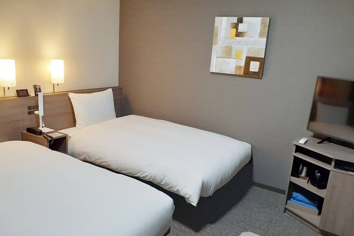 Ein Hotelzimmer mit zwei Betten, einem Nachttisch mit Lampe, einem Fernseher und einem an der Wand hängenden Kunstwerk.