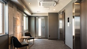 A hallway with a carpeted floor, a chair, a small table, and an elevator. - Hotel Route Inn Matsuyama Chuo -Katsuyama Dori- (Matsuyama)