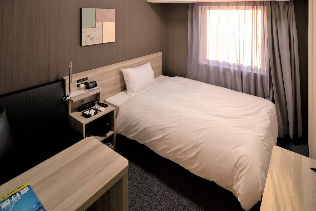Quarto de hotel com cama, mesa de cabeceira, escrivaninha e televisão.